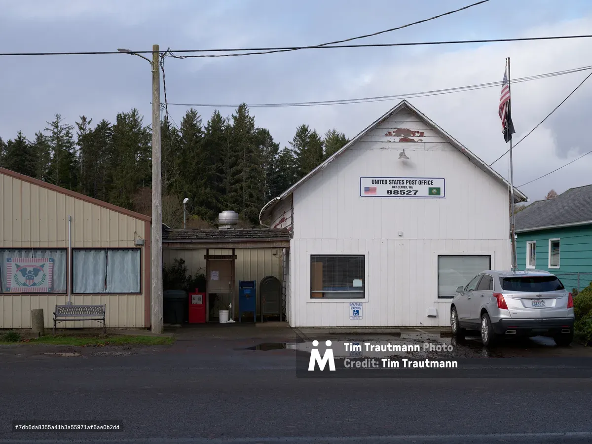 A modest white clapboard United States Post Office building sits along Main Street in Bay Center, Washington, displaying zip code 98527 on its weathered facade. The overcast Pacific Northwest sky casts even light across the scene, where power lines stretch overhead and a silver SUV is parked beside the small-town postal facility. Dense evergreen forests create a dark backdrop behind the building, while neighboring structures including a turquoise house hint at the coastal community's unpretentious character. Puddles on the wet pavement reflect the moody atmosphere of this remote Washington coastal settlement.