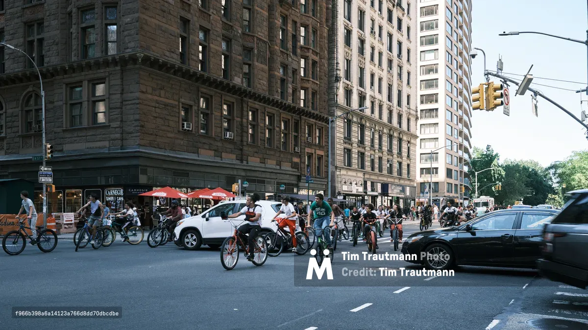 A vibrant street scene captures dozens of cyclists congregating on 7th Avenue in Manhattan, creating an impromptu gathering that transforms the urban thoroughfare into a celebration of bicycle culture. The warm afternoon light bathes the historic brownstone facades and creates long shadows across the asphalt, while modern vehicles navigate around the cycling enthusiasts. The composition juxtaposes New York's architectural heritage—from weathered sandstone buildings to contemporary high-rises—with the grassroots energy of the city's cycling community.