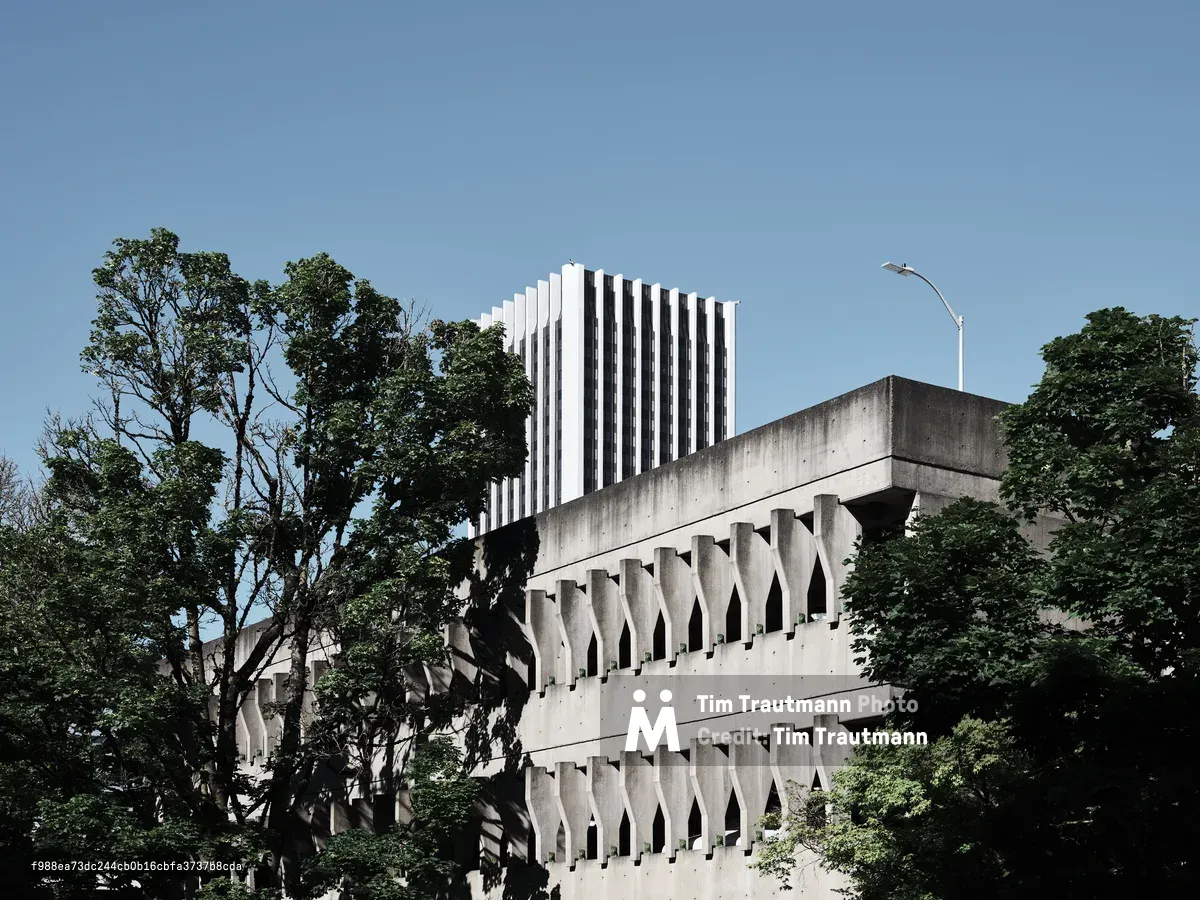 A striking example of Brutalist architecture at Portland State University in downtown Portland, Oregon, featuring a concrete building with repetitive arched openings and geometric patterns. The raw concrete structure is framed by lush green trees under a clear blue sky, with a modern white high-rise building visible in the background.