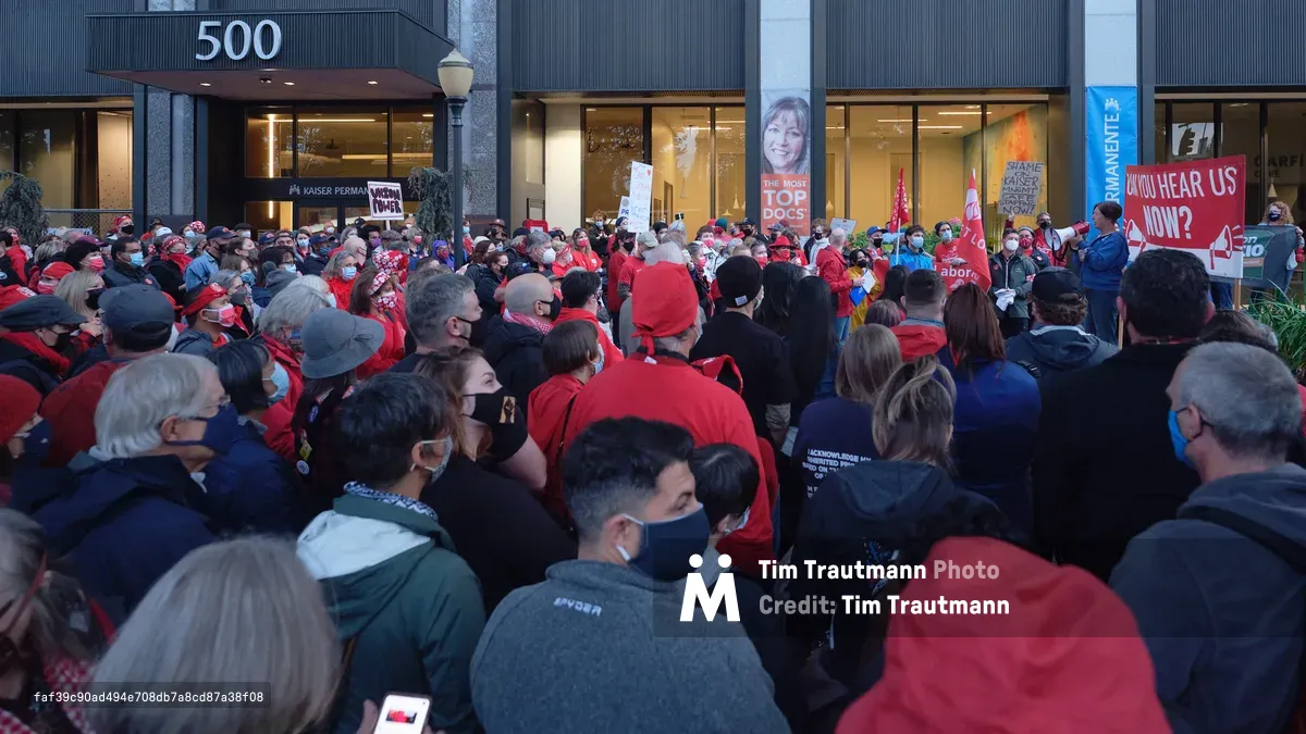 A dense crowd of healthcare workers, predominantly wearing red jackets and face masks, congregates outside the Kaiser Permanente Tower at 500 Northeast Multnomah Street in Portland's Lloyd District. The sea of protesters fills the frame from foreground to the building's glass facade, creating a powerful visual of collective action. Protest signs punctuate the crowd, including one reading 'CAN YOU HEAR US NOW?' while the building's modernist architecture looms behind them, its illuminated interior contrasting with the gathering dusk outside.