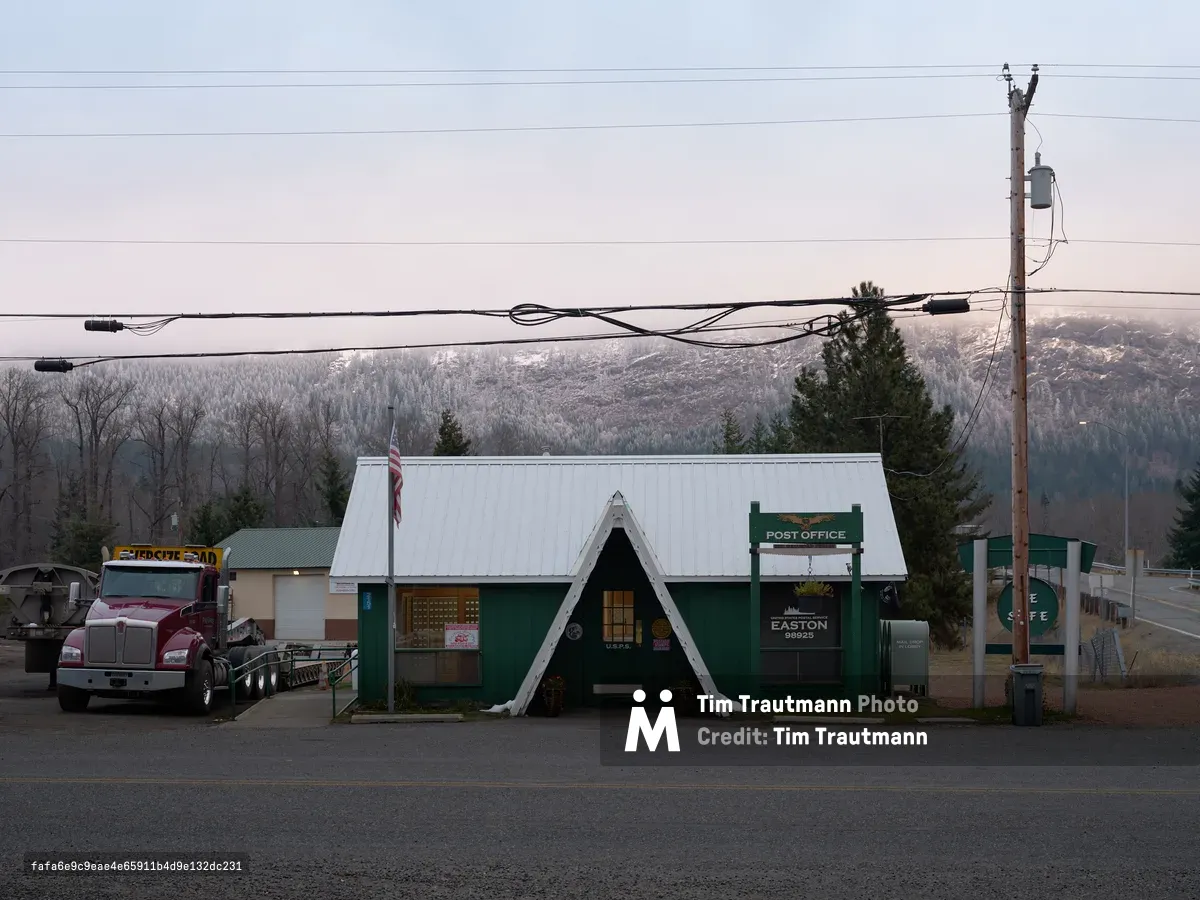 The modest United States Post Office stands sentinel along Cabin Creek Road in Easton, Washington, its distinctive green facade and white A-frame entrance creating an iconic rural postal outpost. Power lines slice through the pale winter sky above, while snow-dusted Cascade Mountain foothills provide a dramatic backdrop to this small-town institution. A logging truck idles nearby, capturing the working character of this Kittitas County community nestled in the mountain pass.