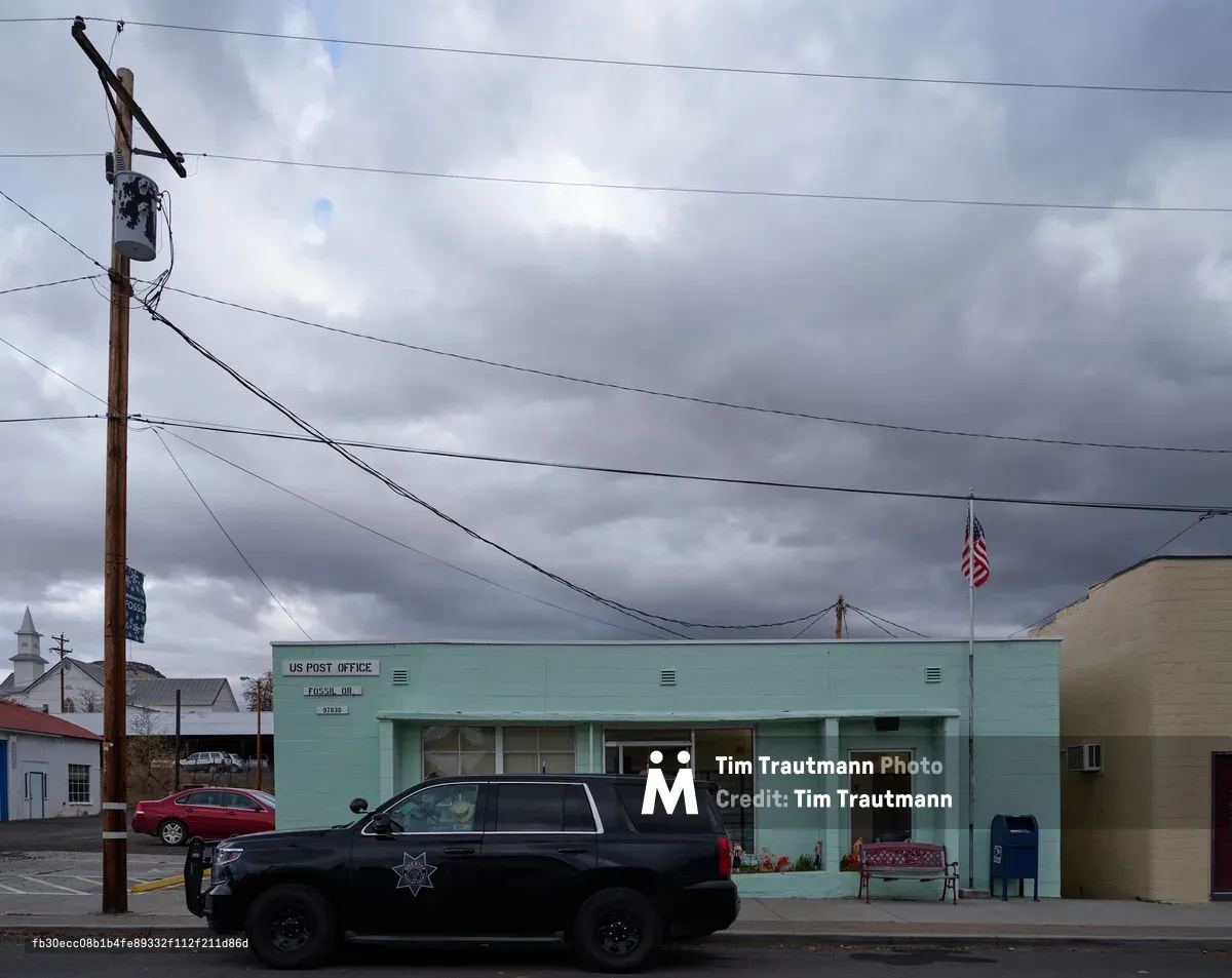 A mint-green United States Post Office stands as the civic anchor of Fossil, Oregon's Main Street, its mid-century modern facade weathered by high desert winds. Ominous gray clouds gather overhead, casting moody light across the utilitarian building while power lines create geometric patterns against the turbulent sky. A black sheriff's SUV and burgundy sedan rest in front of the building, emphasizing the quiet rhythm of small-town life in Wheeler County's rural landscape.