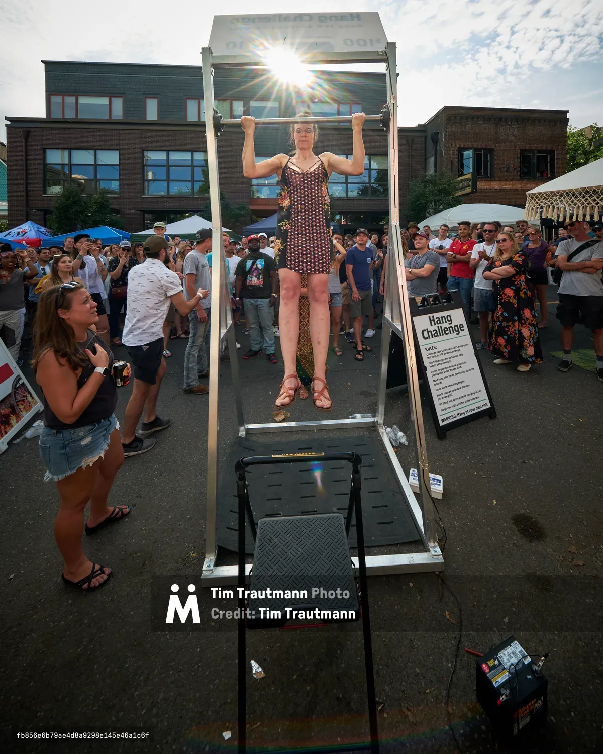 Against the backdrop of Portland's Mississippi Avenue's characteristic modern architecture, a woman in a patterned dress demonstrates remarkable upper body strength during the Hang Challenge at the annual street fair. The bright afternoon sun creates a dramatic starburst effect as it backlights the scene, while dozens of festival-goers gather around the metal hanging apparatus, their attention focused on the athletic feat. The urban setting, complete with brick and contemporary buildings, frames this moment of community celebration and physical prowess in North Portland's vibrant cultural district.