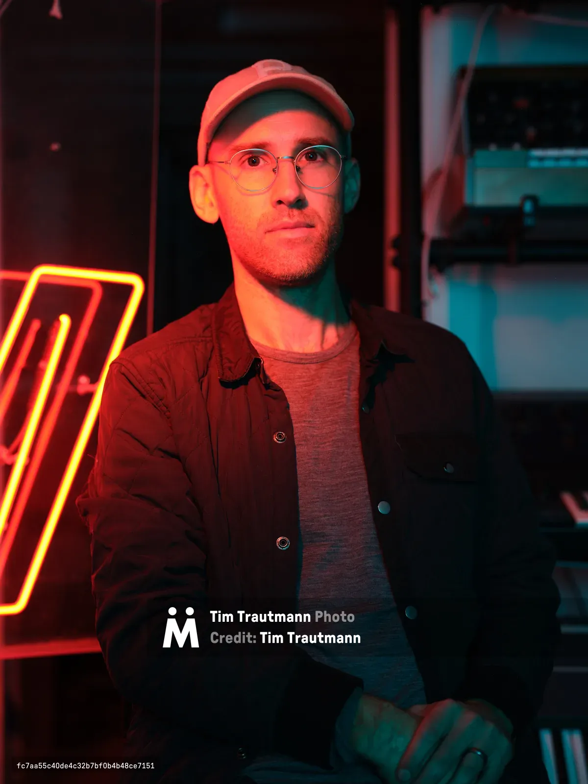 A man wearing glasses, a cap, and layered clothing poses in a dimly music studio with vibrant red and blue neon lighting in the background. The atmospheric lighting creates a moody scene of a music studio.