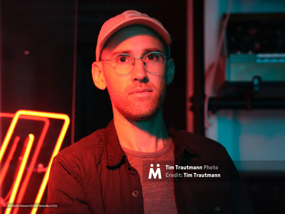 A young man wearing round glasses and a baseball cap poses in front of colorful neon lighting in what appears to be a music studio. The dramatic lighting creates red and blue tones across his face and casual button-up shirt.