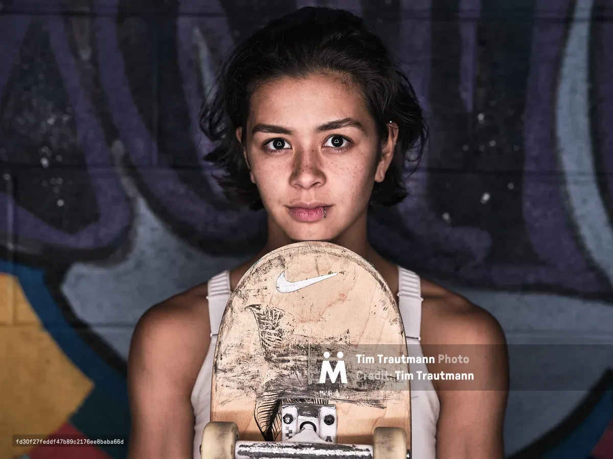 A close-up portrait of pro skateboarder Christiana Means holding a well-worn Nike skateboard deck up to her chin, gazing directly into the camera with a calm, focused expression. She has short dark hair, freckles, and a small lip piercing, and wears a white tank top. A colorful graffiti mural in purple, blue, and yellow serves as the background.