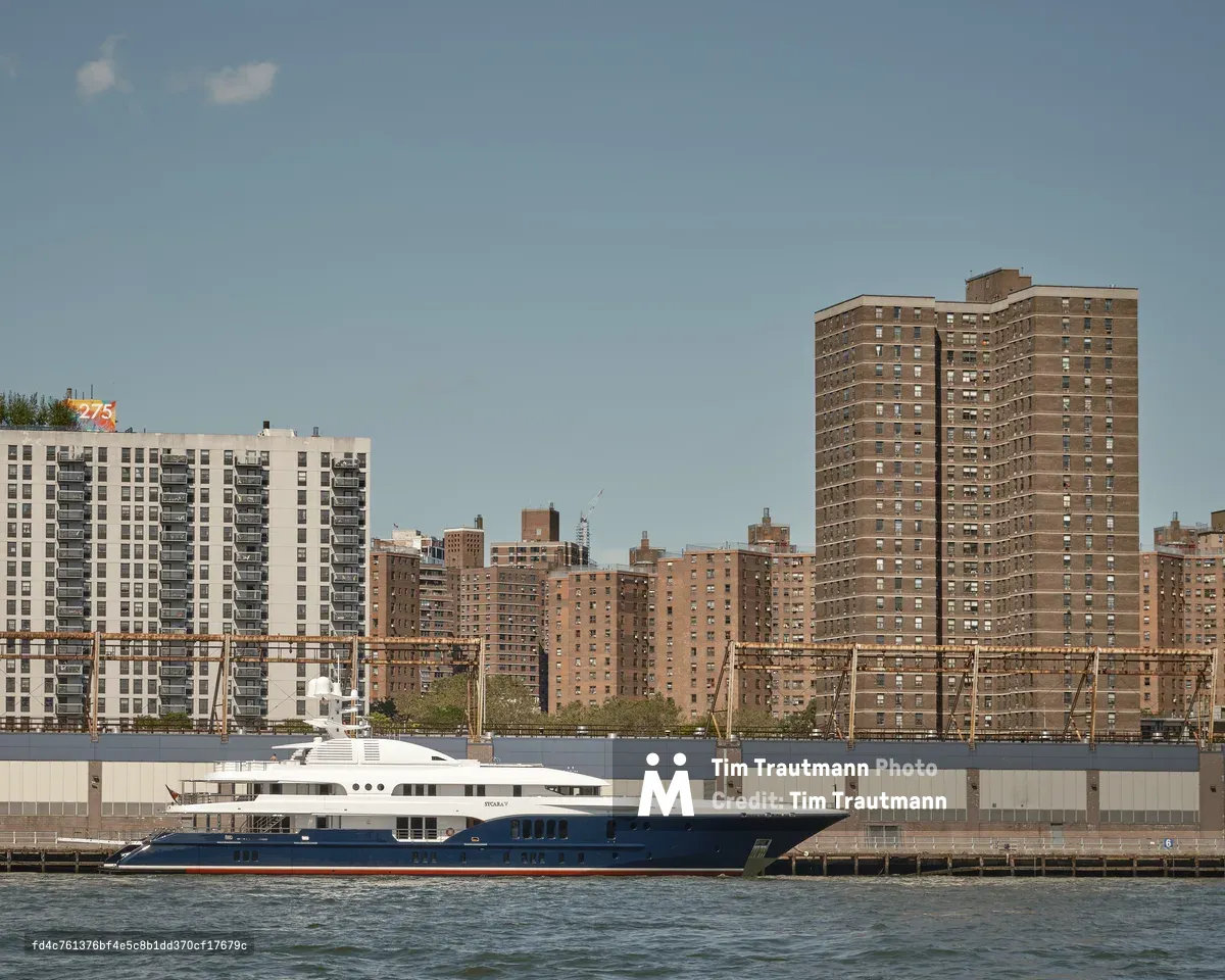 A large luxury superyacht with a deep navy blue hull, red waterline stripe, and white superstructure is moored at a pier along the Manhattan waterfront, photographed from across the East River in Brooklyn. The vessel's name appears to read "Sycara V" on the hull. Behind the yacht, rusted industrial pier infrastructure and a large grey waterfront building line the shore. Rising above are multiple massive brown brick mid-rise residential housing towers characteristic of Lower East Side public housing complexes. A colorful rooftop mural bearing the number "275" is visible on one of the shorter buildings at left. The sky is clear and hazy blue, with a single small cloud. The contrast between the opulent private yacht and the dense public housing towers behind it is stark.