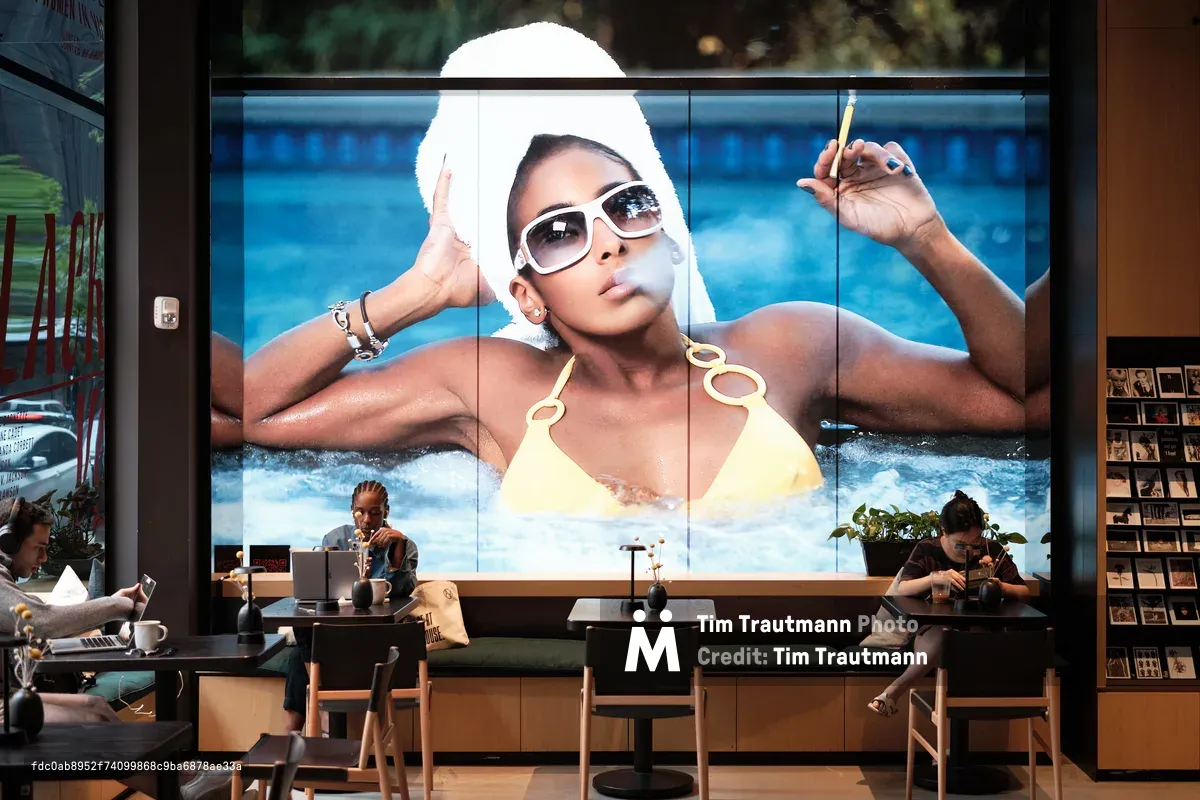 Within the contemporary walls of Fotografiska New York, patrons sip coffee beneath a towering advertisement featuring a glamorous woman in oversized sunglasses and a white head wrap, luxuriating poolside in a yellow bikini. The juxtaposition creates a striking dialogue between commercial imagery and intimate café culture, where warm wooden furnishings and soft natural light filtering through floor-to-ceiling windows provide an elegant counterpoint to the bold, sun-drenched advertisement above. Two visitors occupy the space quietly, one reading at a corner table while another works at her laptop, seemingly oblivious to the larger-than-life presence watching over their mundane moments.