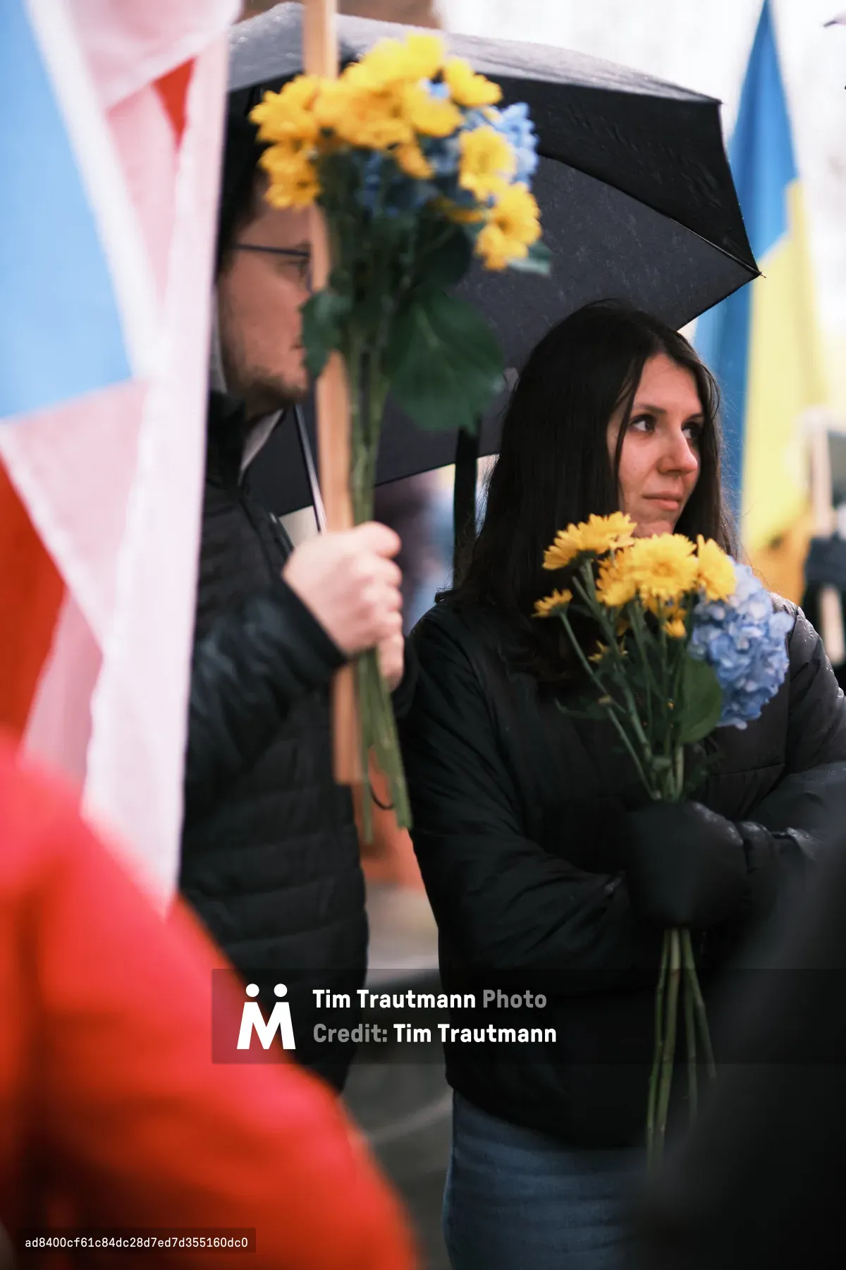 A solemn woman clutches yellow chrysanthemums and blue hydrangeas close to her chest while standing beneath a black umbrella held by a companion. The scene unfolds against a backdrop of Ukrainian flag colors—vibrant blue and yellow banners creating a patriotic tapestry behind the protesters. Her contemplative gaze and the protective gesture of holding flowers suggest both defiance and mourning, captured in the soft, overcast light outside Portland's Revolution Hall during an anti-war demonstration.