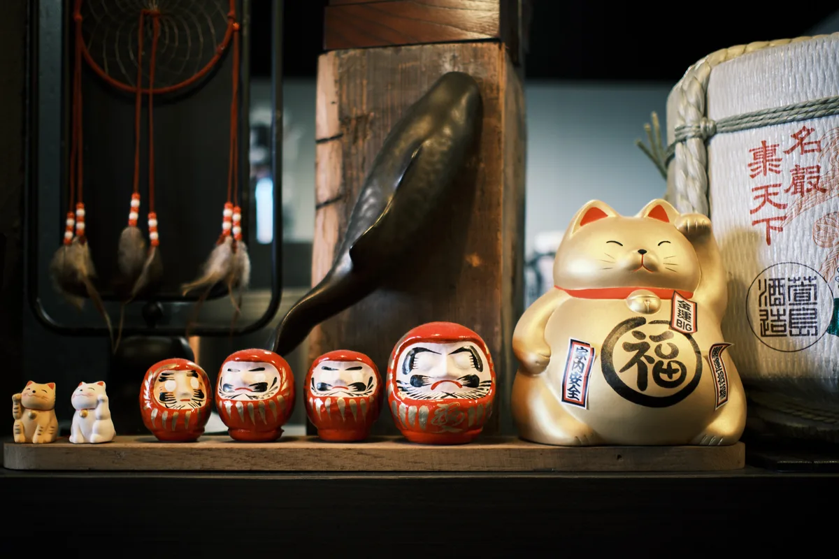 A curated arrangement of Japanese good luck talismans creates an intimate shrine within Portland's Sushi Mazi restaurant. Four vermillion daruma dolls sit in perfect progression alongside a golden maneki-neko cat, while traditional rope-bound sake containers and ceramic vessels frame the composition in warm, ambient lighting. The weathered wooden shelf and shadowy depths behind speak to authenticity and reverence, transforming everyday restaurant décor into a meditation on prosperity and protection.