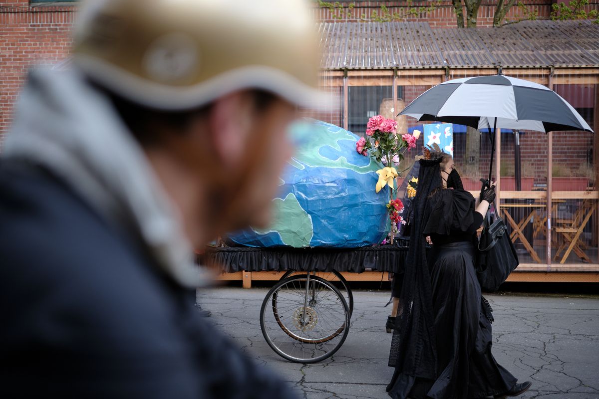 A solemn funeral scene in Portland, Oregon showing mourners in black gathered around an ornate horse-drawn hearse decorated with blue fabric and flowers, with an umbrella providing shelter during what appears to be a rainy day.