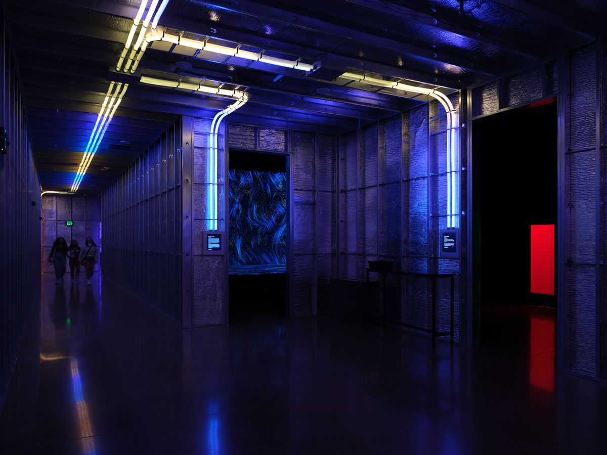 A dramatic interior space featuring blue LED lighting, exposed ceiling infrastructure, reflective floors, and modern architectural elements with silhouetted figures in the background. The space showcases contemporary design with neon accent lighting and industrial materials.
