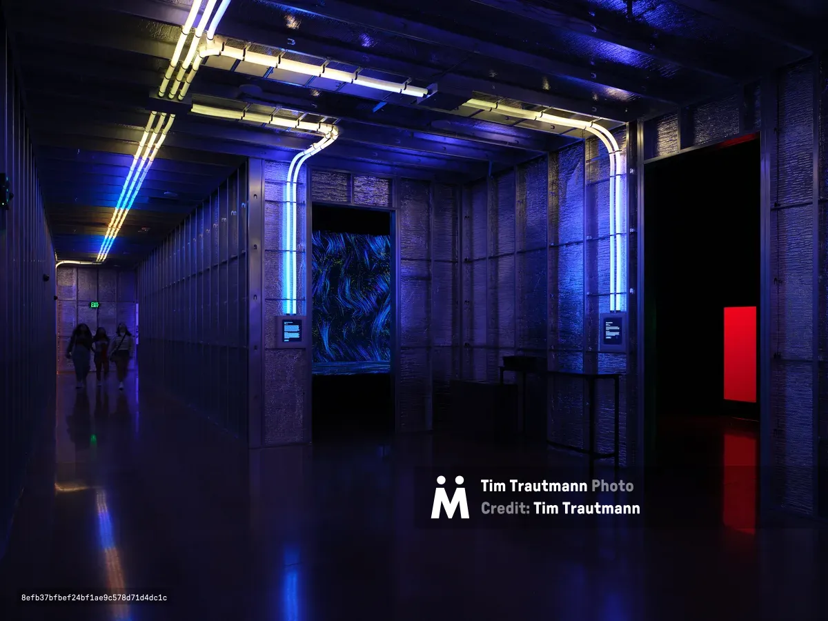 A dramatic interior space featuring blue LED lighting, exposed ceiling infrastructure, reflective floors, and modern architectural elements with silhouetted figures in the background. The space showcases contemporary design with neon accent lighting and industrial materials.