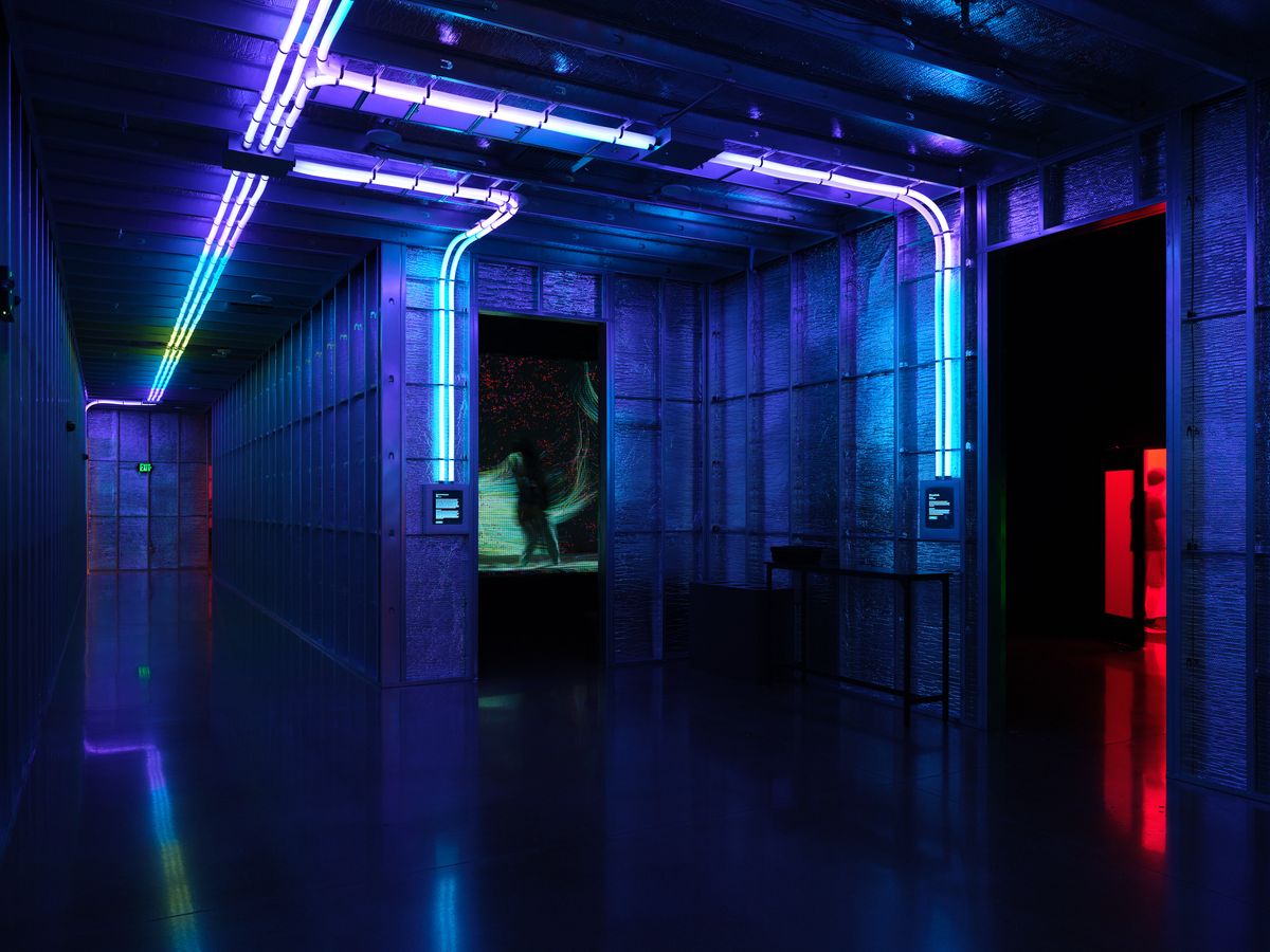 A dimly lit interior space with dramatic blue and purple neon lighting creating a futuristic atmosphere. The industrial-style room features concrete walls, exposed ceiling elements, and curved neon light installations that reflect on the polished floor, with accent lighting in red and green visible in the background.