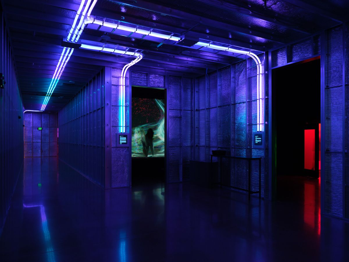 A modern gallery interior bathed in dramatic blue and purple neon lighting, featuring curved neon tubes along concrete walls and ceiling, with a reflective floor and an illuminated display screen visible in the background. The space has an industrial, cyberpunk aesthetic with clean architectural lines and atmospheric lighting creating a futuristic ambiance.