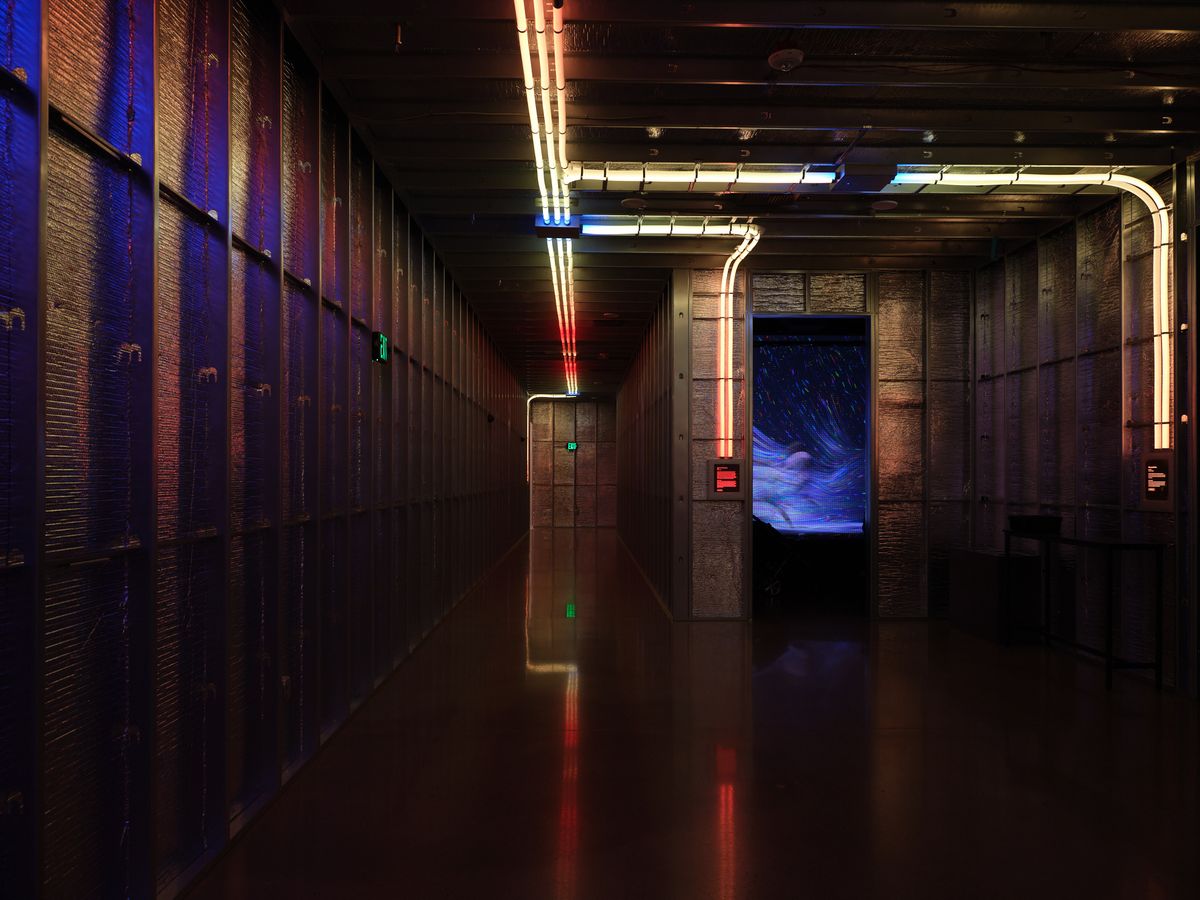 A dramatic interior hallway featuring industrial brick walls, neon tube lighting in red and blue colors, reflective floors, and a large blue digital screen at the end of the corridor. The space has a cyberpunk aesthetic with modern architectural elements and atmospheric lighting.