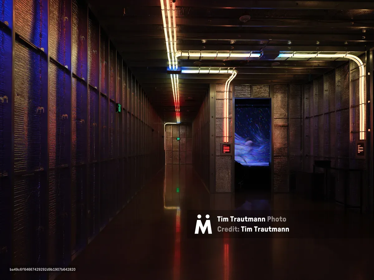 A dramatic interior hallway featuring industrial brick walls, neon tube lighting in red and blue colors, reflective floors, and a large blue digital screen at the end of the corridor. The space has a cyberpunk aesthetic with modern architectural elements and atmospheric lighting.
