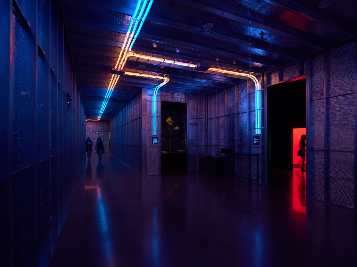 A dramatic interior corridor featuring striking blue and orange neon lighting strips along the ceiling and walls, creating a cyberpunk atmosphere with silhouetted figures walking through the illuminated space. The modern architectural design includes exposed ceiling elements and reflective flooring that amplifies the colorful LED lighting effects.