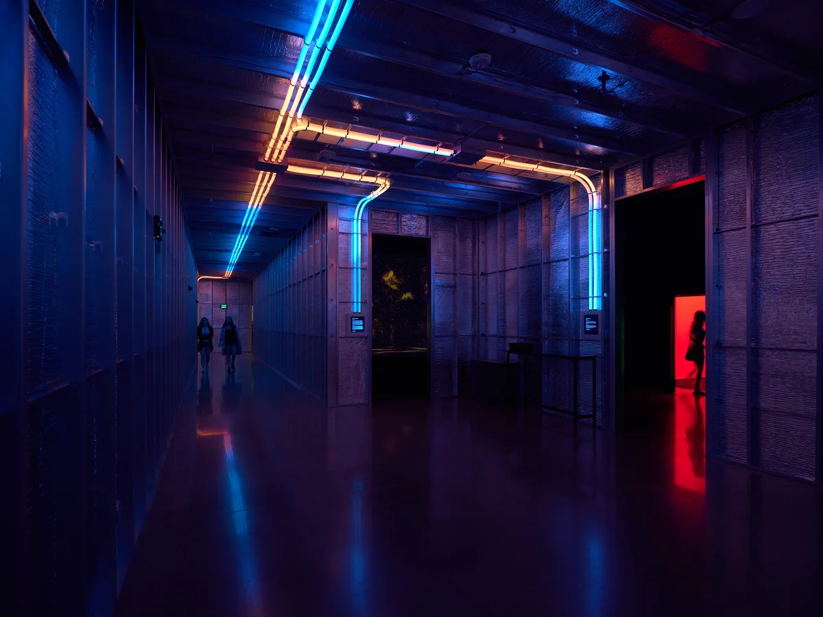 A dramatic interior corridor featuring striking blue and orange neon lighting strips along the ceiling and walls, creating a cyberpunk atmosphere with silhouetted figures walking through the illuminated space. The modern architectural design includes exposed ceiling elements and reflective flooring that amplifies the colorful LED lighting effects.