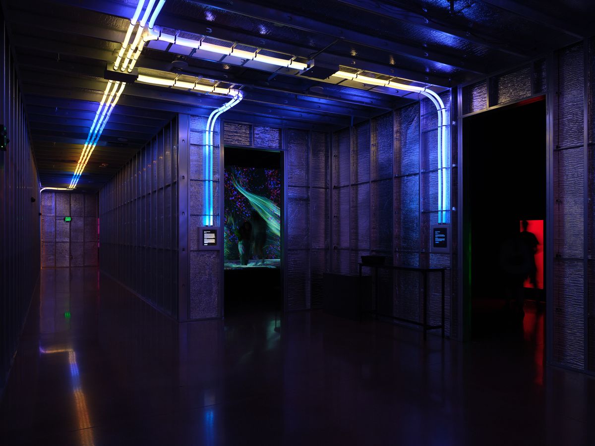 A dramatic interior hallway at Hopscotch in Portland, Oregon features industrial architecture with exposed brick walls, neon blue and purple lighting strips running along the ceiling and walls, creating a cyberpunk atmosphere. The corridor leads to what appears to be an entertainment area with glowing screens and digital displays visible in the distance.