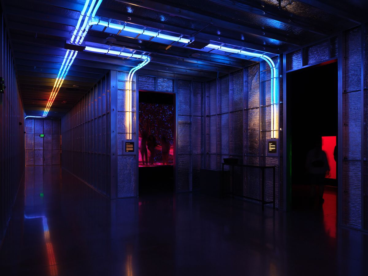 A modern gallery interior at Hopscotch in Portland, Oregon featuring dramatic blue and red neon lighting strips running along the ceiling and walls, creating a sci-fi atmosphere with reflective floors and industrial architecture.