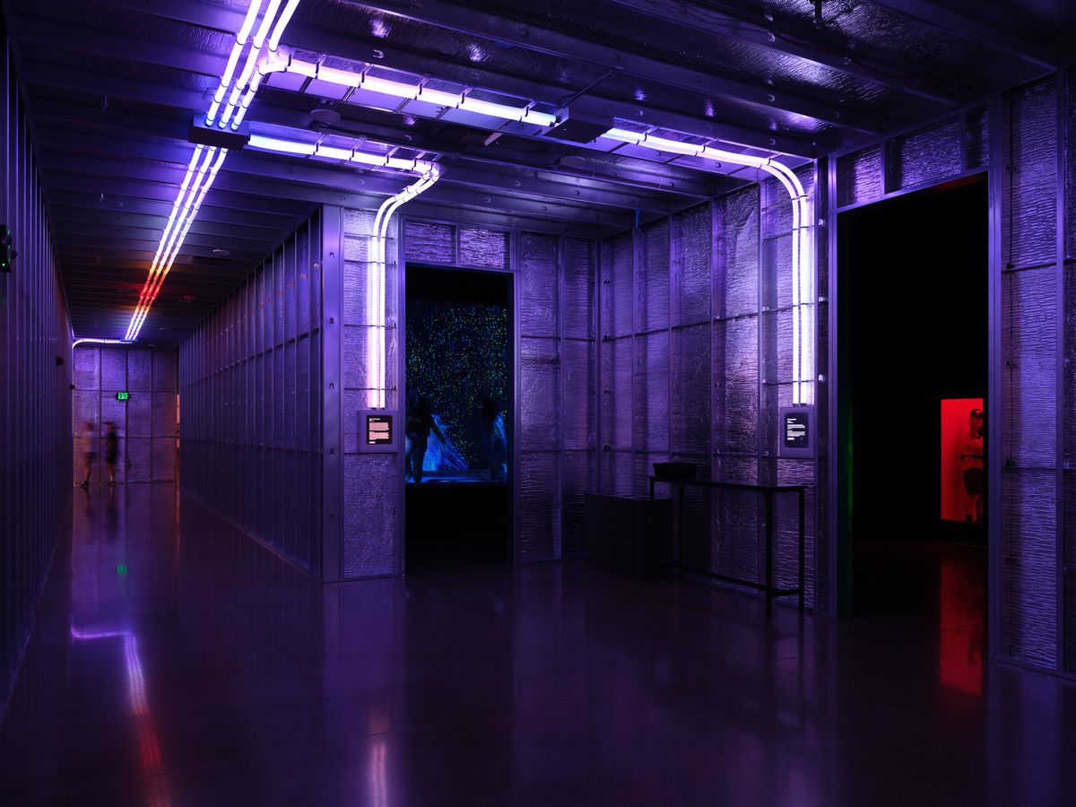 A dramatic interior space at Hopscotch in Portland, Oregon featuring purple and blue neon lighting, concrete walls, and reflective floors creating a cyberpunk-inspired atmosphere. The space includes curved neon tubes on the ceiling, display screens, and red accent lighting along dark corridors.