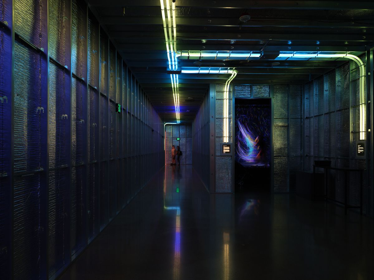 A dimly lit industrial corridor featuring concrete walls, exposed pipes, and vibrant neon lighting in blues and yellows. The long hallway has a wet reflective floor and atmospheric lighting that creates a cyberpunk aesthetic.