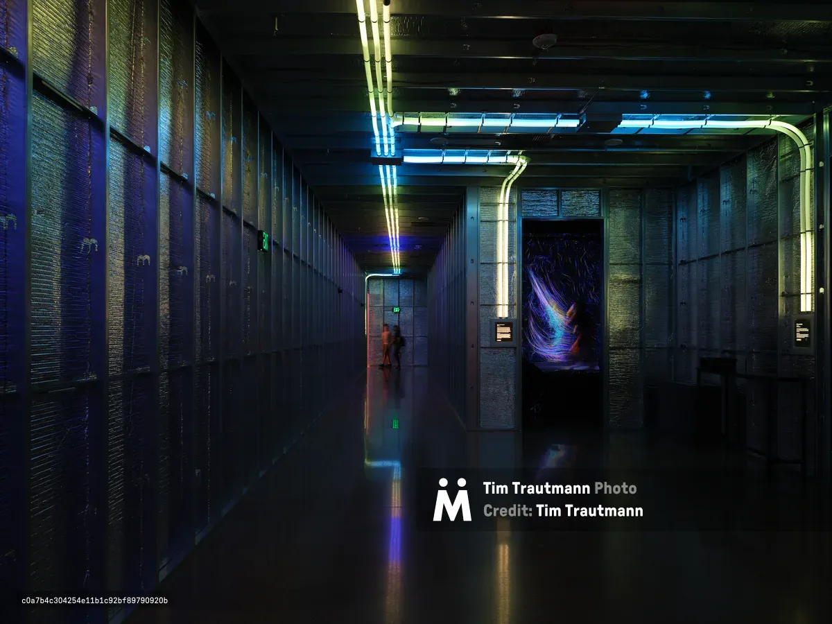 A dimly lit industrial corridor featuring concrete walls, exposed pipes, and vibrant neon lighting in blues and yellows. The long hallway has a wet reflective floor and atmospheric lighting that creates a cyberpunk aesthetic.