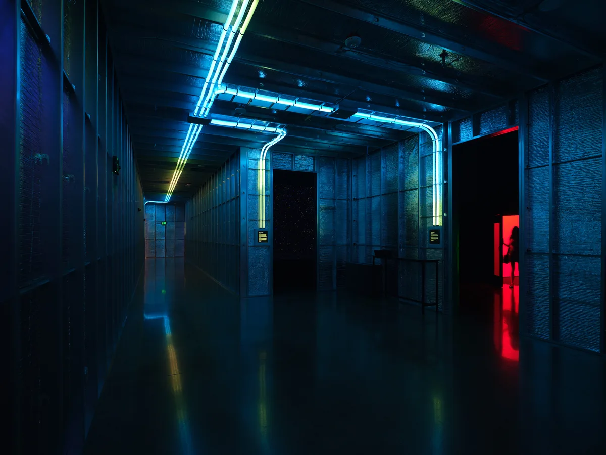 A dramatic underground corridor featuring flooded concrete floors that reflect vibrant blue and red neon lighting from overhead fixtures. The industrial space has exposed pipes and ventilation systems along the ceiling, creating a cyberpunk aesthetic with strong architectural lines leading into darkness.