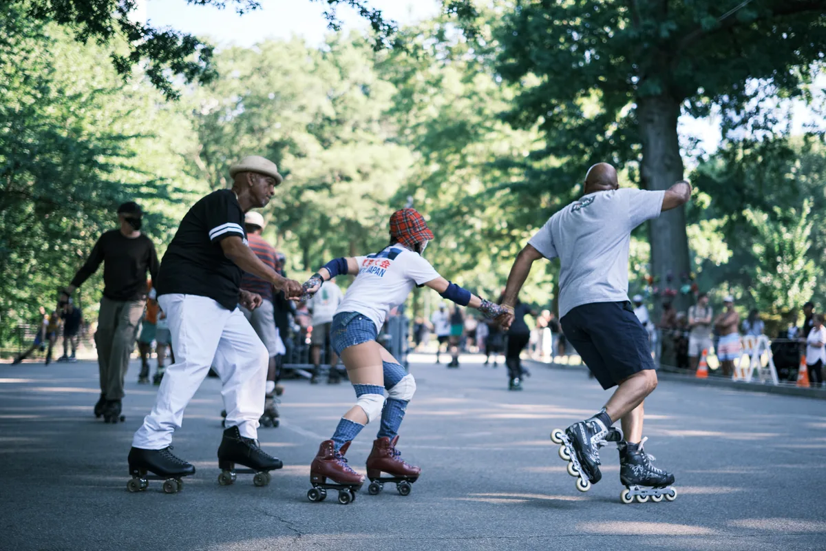 Three generations converge in a spirited tug-of-war beneath Central Park's dappled canopy, their determination etched in strained postures and interlocked grips. An elderly man in a beret anchors one end while a young participant in roller skates and tartan provides the middle link, connected to a man in inline skates who leans into the effort. Filtered sunlight streams through the verdant foliage, casting dancing shadows across the park's asphalt pathway where weekend recreation unfolds.
