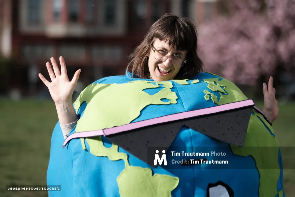 A jubilant woman wearing wire-rimmed glasses emerges from an oversized Earth costume, her arms raised in triumphant celebration against the soft-focus backdrop of Portland's Revolution Hall. The handcrafted globe features vivid azure oceans and lime-green continents, while a distinctive pink laptop computer sits incongruously across the equator. Golden afternoon light illuminates her genuine smile and the costume's felt textures, creating an atmosphere of playful environmental activism in Oregon's Central Eastside district.
