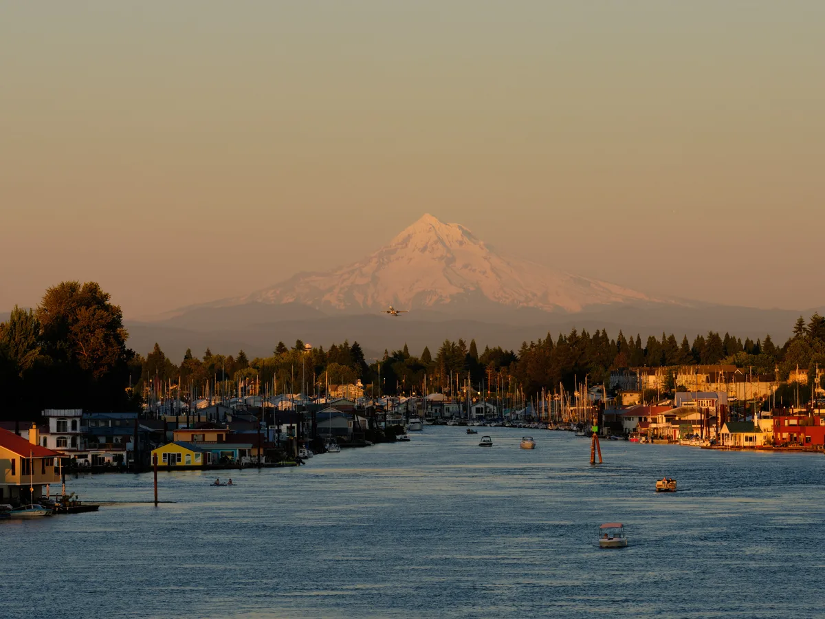 Mount Hood rises majestically through the amber-tinted atmosphere as golden hour light bathes Portland's North Harbor along the Columbia River. The snow-capped volcanic peak provides a dramatic backdrop to the bustling waterfront community, where colorful houseboats and maritime structures line the calm waters near Portland International Airport. Small aircraft can be seen ascending through the warm, hazy sky, while pleasure boats dot the serene river channel that flows between forested shores.
