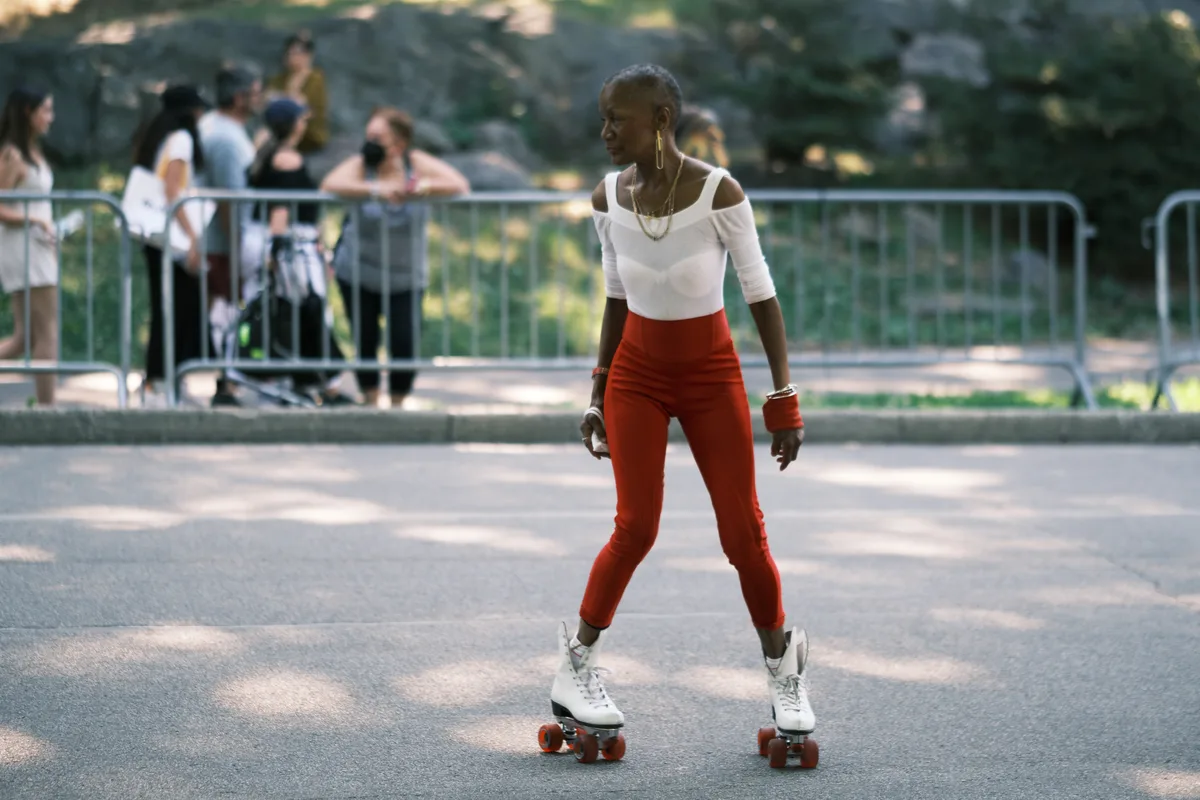 A woman glides gracefully on vintage white roller skates across the sun-dappled asphalt of Central Park's skating rink. She wears a striking ensemble of vermillion high-waisted pants and a crisp white cold-shoulder top, her movements captured mid-stride as golden hour light filters through the leafy canopy above. Behind her, metal barriers frame other park visitors who blur into the dreamy bokeh of late afternoon shadows and light. The scene pulses with nostalgic energy and urban recreation.