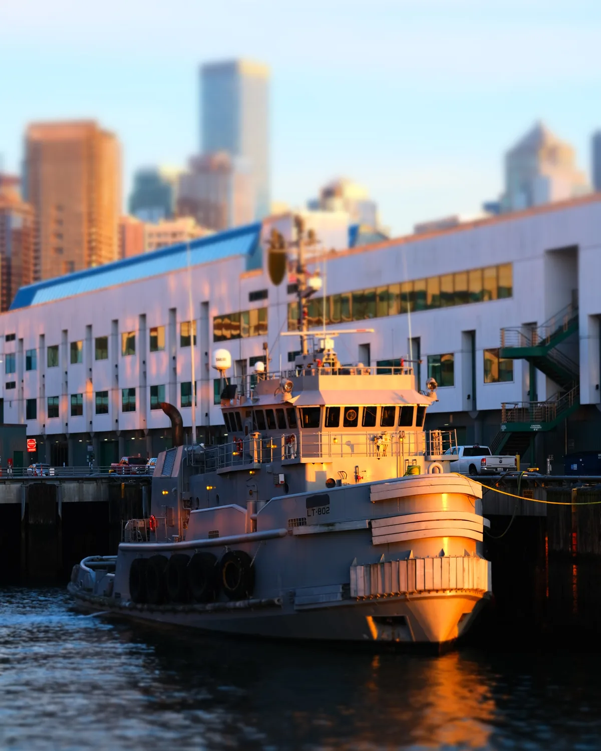 A working tugboat designated T-802 basks in the amber light of golden hour at Seattle's Pier 70, its white hull and wheelhouse glowing warmly against Elliott Bay's dark waters. The vessel's utilitarian design and illuminated cabin windows suggest the quiet end of a working day, while the soft-focused skyline of downtown Seattle rises majestically in the background. The modern waterfront development creates a striking architectural contrast to the honest functionality of the maritime workboat, capturing the intersection of Seattle's industrial heritage and urban evolution.
