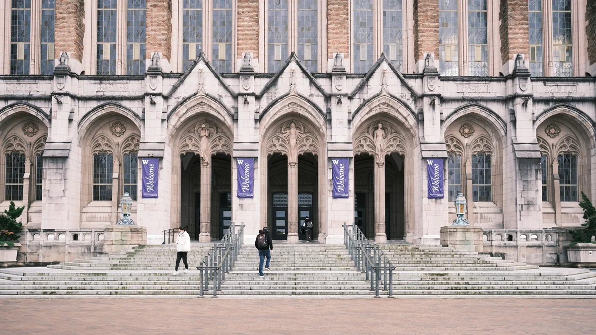 The imposing Gothic Revival facade of Suzzallo Library dominates the frame with its soaring pointed arches and ornate stonework, crowned by rows of tall mullioned windows that catch the soft daylight. Purple welcome banners flutter between the carved columns, while two solitary figures navigate the broad stone steps, dwarfed by the architectural monumentality. The warm-toned limestone contrasts beautifully with the deep shadows cast by the elaborate tracery, creating a scene that balances academic solemnity with accessible human scale.