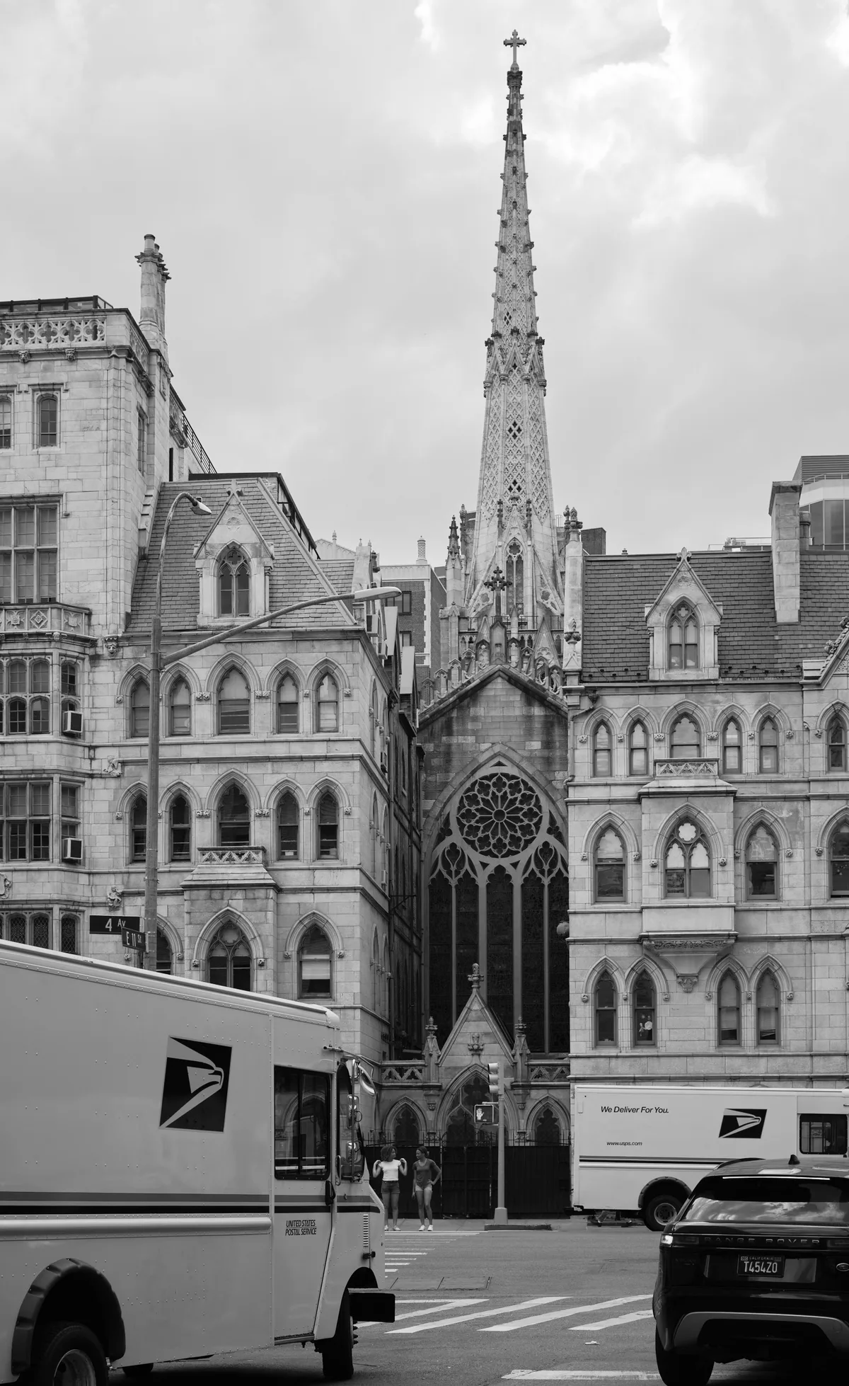 Grace Church's ornate Gothic Revival spire soars majestically above the bustling streets of Greenwich Village, its intricate stonework and cross-topped pinnacle creating a dramatic vertical counterpoint to the surrounding historic buildings. The black and white composition emphasizes the architectural details of the church's elaborate facade, complete with rose window and pointed arches, while postal trucks and urban activity ground the sacred structure in contemporary New York life. Soft, overcast lighting bathes the limestone surfaces in gentle gradations of gray, highlighting the craftsmanship of this 1840s ecclesiastical masterpiece.