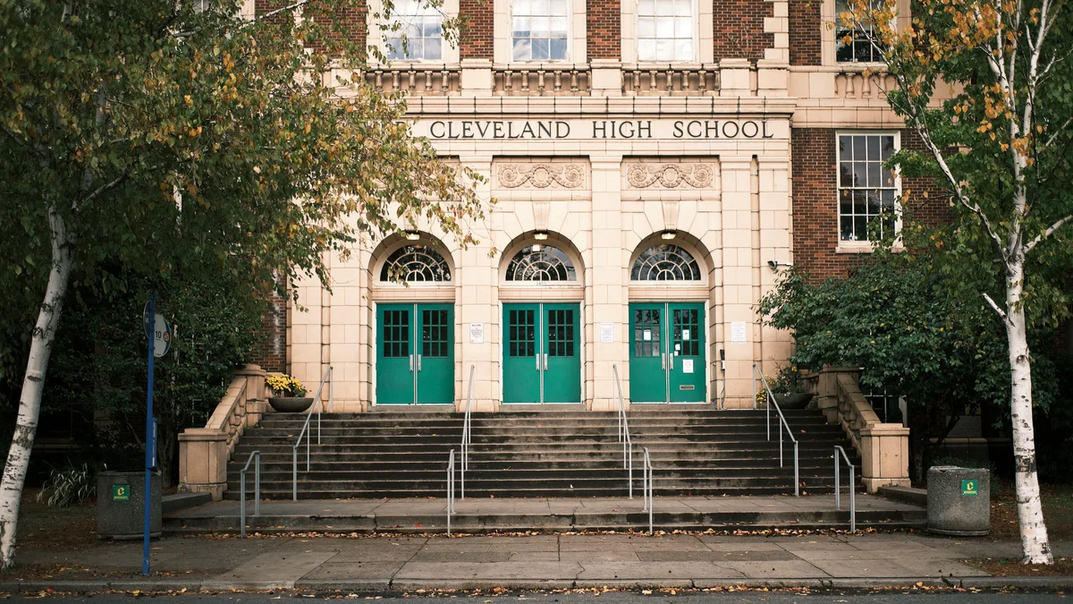 The stately historic facade of Grover Cleveland High School in Portland, Oregon, photographed on an autumn day. The building features a classical red brick and cream stone exterior with three arched entryways fitted with teal green double doors and fanlight windows above. "Cleveland High School" is carved in stone above the central entrance. Wide stone steps with metal handrails lead up to the three entrances. White-barked birch trees with yellowing autumn leaves frame the building on both sides, and fallen leaves scatter the sidewalk in the foreground.