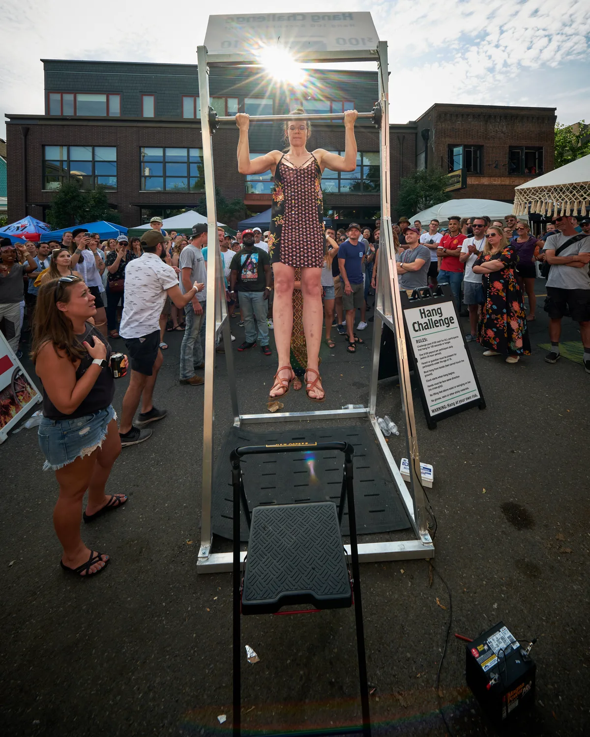 Against the backdrop of Portland's Mississippi Avenue's characteristic modern architecture, a woman in a patterned dress demonstrates remarkable upper body strength during the Hang Challenge at the annual street fair. The bright afternoon sun creates a dramatic starburst effect as it backlights the scene, while dozens of festival-goers gather around the metal hanging apparatus, their attention focused on the athletic feat. The urban setting, complete with brick and contemporary buildings, frames this moment of community celebration and physical prowess in North Portland's vibrant cultural district.
