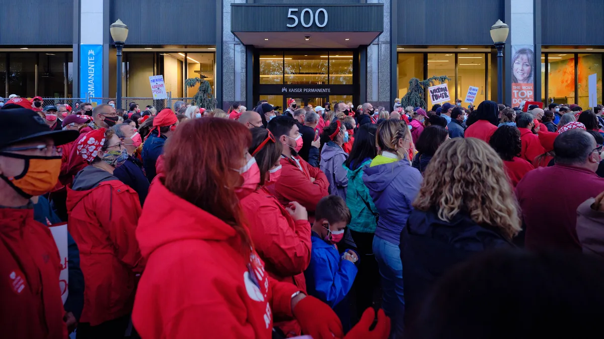 A dense crowd of healthcare workers, predominantly wearing red Kaiser Permanente scrubs and winter jackets, gathers in solidarity outside the Kaiser Permanente Tower at 500 Northeast Multnomah Street in Portland's Lloyd District. The sea of crimson creates a powerful visual statement against the modern glass and steel facade of the medical facility, while protest signs punctuate the crowd and evening light casts a determined atmosphere over the assembled nurses. Face masks worn by many participants underscore the contemporary healthcare context of their labor action.