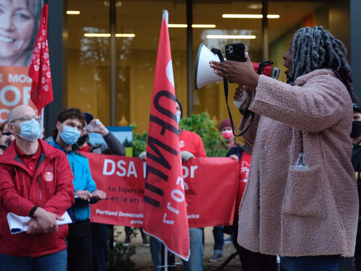 A passionate healthcare advocate with distinctive gray locs commands attention through a white megaphone, her voice cutting through the urban afternoon air outside Kaiser Permanente's imposing glass tower in Portland's Lloyd District. Behind her, masked nurses in crimson jackets clutch red DSA banners, their determined faces illuminated by the warm fluorescent glow spilling from the corporate lobby. The scene pulses with collective energy as workers prepare for their impending strike, their unified presence creating a stark contrast against the sterile corporate backdrop.
