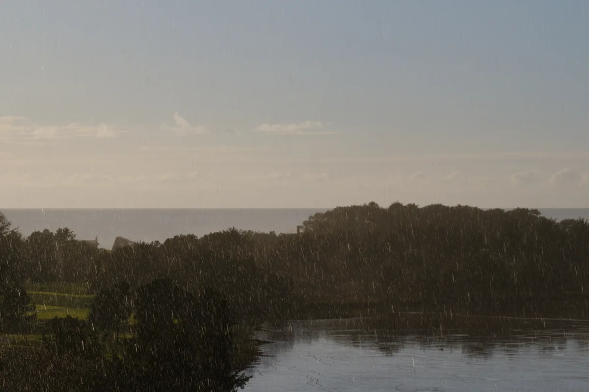 A dramatic scene of intense rainfall falling over a forested coastal landscape in Gleneden Beach, Oregon, with visible rain streaks across the frame, dark storm clouds overhead, and a body of water in the foreground reflecting the moody sky.