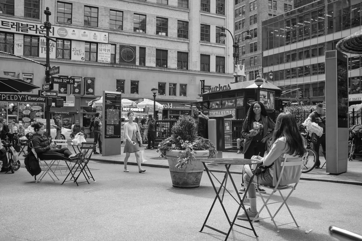 In the bustling heart of Herald Square, Manhattan, pedestrians claim moments of urban tranquility at scattered café tables amid the commercial chaos. The monochromatic composition captures the rhythmic dance of city life, where solitary figures pause with coffee and conversation against a backdrop of towering brick facades and Korean signage. Geometric folding chairs and planters create intimate pockets of respite, while streams of shoppers navigate between the outdoor seating and storefront displays.