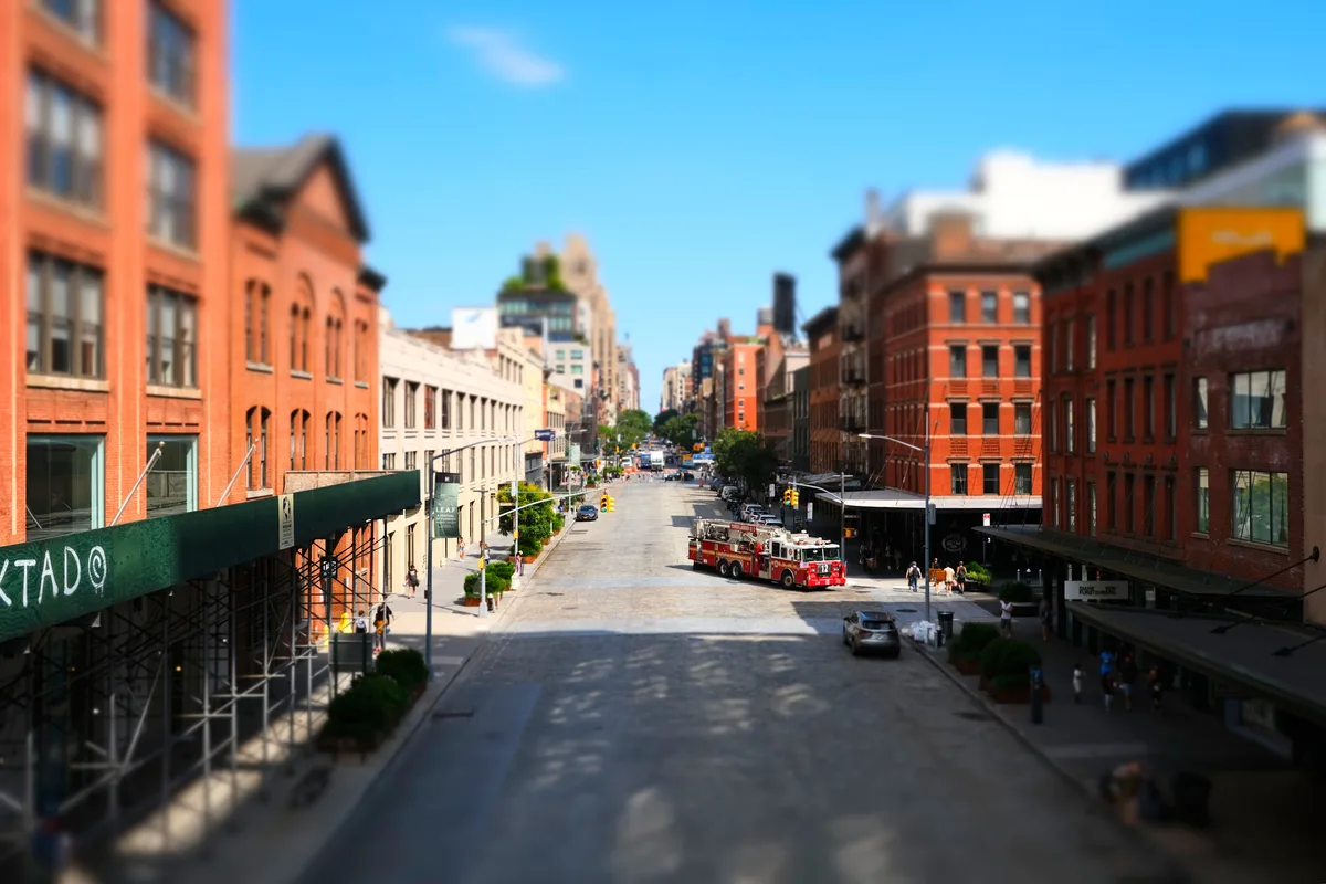 A dramatic tilt-shift perspective captures the intersection of 10th Avenue and West 14th Street from the elevated High Line walkway, where industrial-era brick warehouses frame a sun-drenched corridor. A red fire truck commands the center of the composition, its emergency presence juxtaposed against the leisurely pedestrians exploring this transformed Meatpacking District landscape. The shallow depth of field renders the urban canyon in miniature-like detail, while brilliant blue skies crown the historic neighborhood's rebirth from industrial grit to cultural destination.