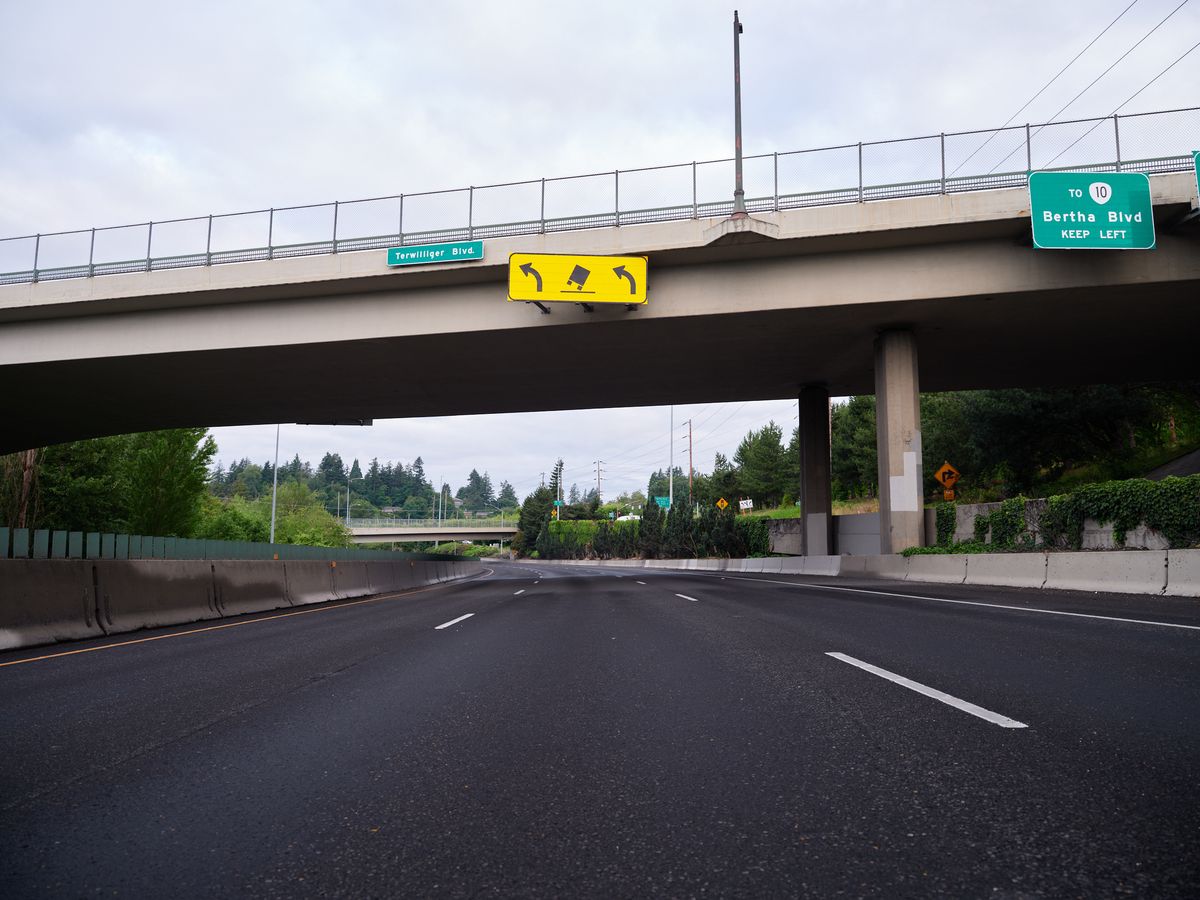 A view from beneath a concrete highway overpass showing Terwilliger Boulevard signage and directional arrows, with the roadway extending into the distance through a forested area in Portland's Hillsdale neighborhood. Green highway signs indicate routes to Bertha Boulevard and Highway 10.