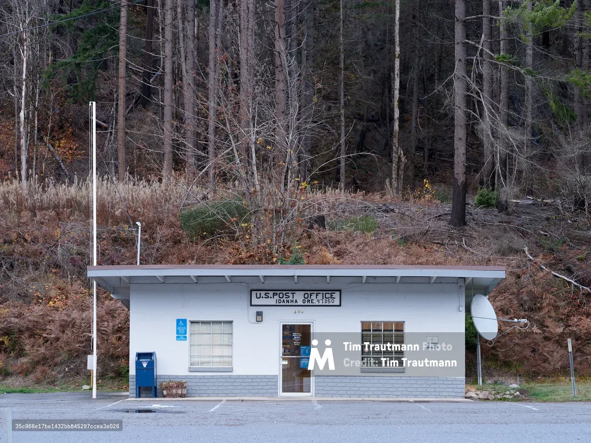 A modest white cinder block post office building sits beneath a hillside of bare deciduous trees and towering evergreens in the small Oregon community of Idanha. The single-story structure, marked with its official USPS designation and zip code 97350, features classic government architecture with a flat roof, flagpole, and satellite dish. Autumn's dormant landscape of rust-colored undergrowth and skeletal branches creates a stark backdrop, emphasizing the building's role as a vital connection point in this remote Cascade foothills settlement along the North Santiam Highway.