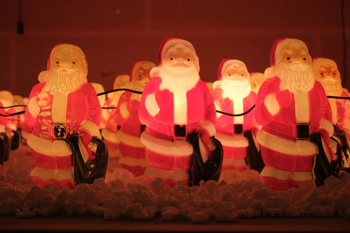 A mesmerizing battalion of identical illuminated Santa Claus figures stands sentinel in Portland's Concordia neighborhood, their warm amber glow piercing the burgundy winter darkness. Each plastic saint clutches a black lantern, creating a rhythmic pattern of jolly faces that stretches into the atmospheric haze. The figures emit an ethereal radiance against the moody evening sky, transforming Northeast Killingsworth Street into a surreal Christmas dreamscape where mass-produced holiday cheer takes on an almost mystical quality.