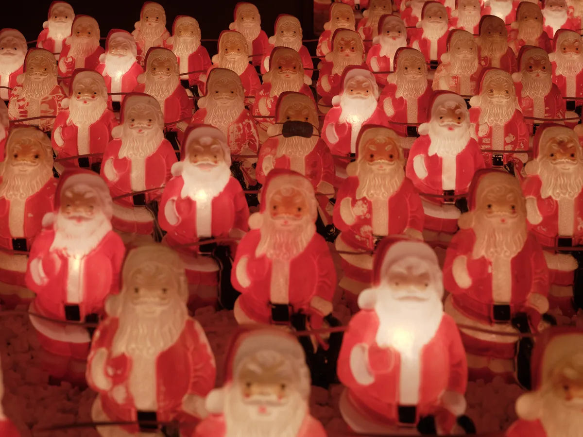Dozens of molded plastic Santa Claus figures glow with warm amber light in Chris Willis's annual art installation at a Sellwood consignment gallery. The repetitive crimson-clad figures create a mesmerizing pattern that stretches into darkness, their identical jolly expressions caught between whimsy and unsettling uniformity. The installation transforms the humble Christmas decoration into a commentary on mass production and holiday commercialization, with each illuminated Santa casting a soft pool of light in the gallery's shadowy interior.