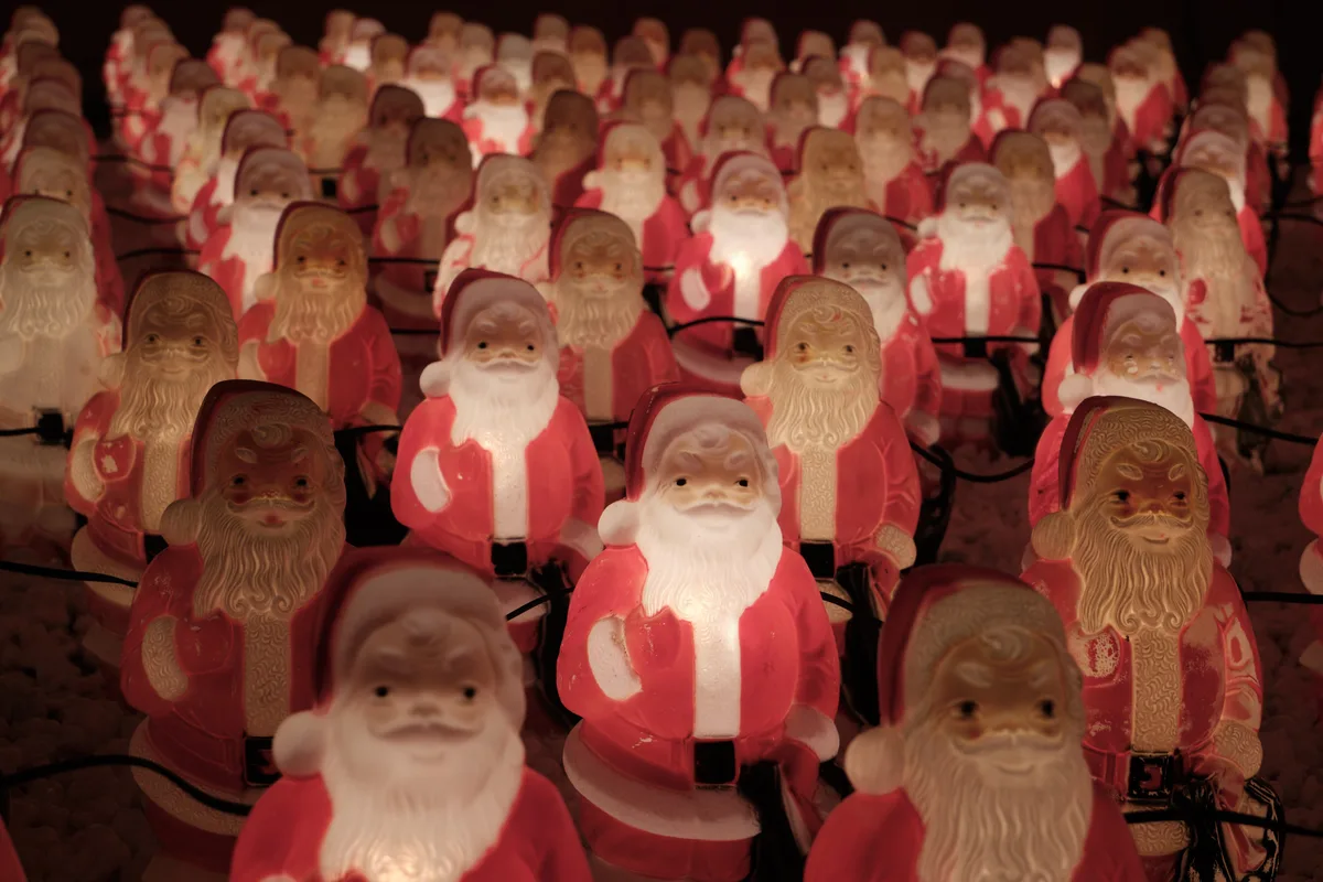 Hundreds of plastic Santa Claus figurines create a mesmerizing sea of red and white beneath the glow of string lights in Portland's Concordia neighborhood. The illuminated collection stretches into darkness, each jolly figure casting gentle shadows while warm light emanates from within their translucent forms. This enchanting holiday installation transforms an ordinary urban space into a whimsical wonderland, where countless identical Santas stand in silent formation like a benevolent Christmas militia.