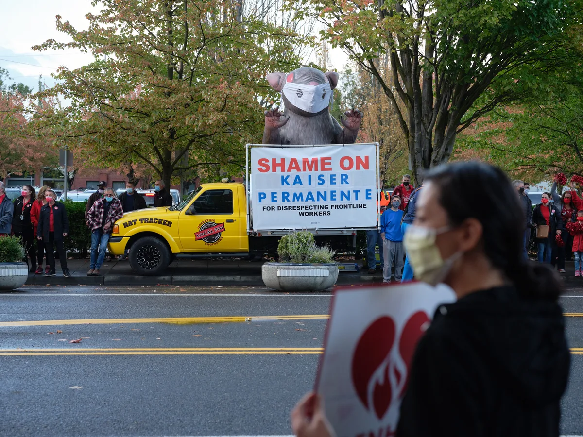 A towering inflatable rat protest figure dominates the scene outside Kaiser Permanente Tower in Portland's Lloyd District, its menacing presence amplified by autumn's golden canopy. Red-clad nurses and supporters gather in solidarity along Northeast Multnomah Street, their masked faces reflecting both pandemic precautions and labor tensions. The bold white protest sign declaring 'SHAME ON KAISER PERMANENTE' cuts through the overcast afternoon light, while a yellow Rat Tracker truck anchors the demonstration with industrial determination.