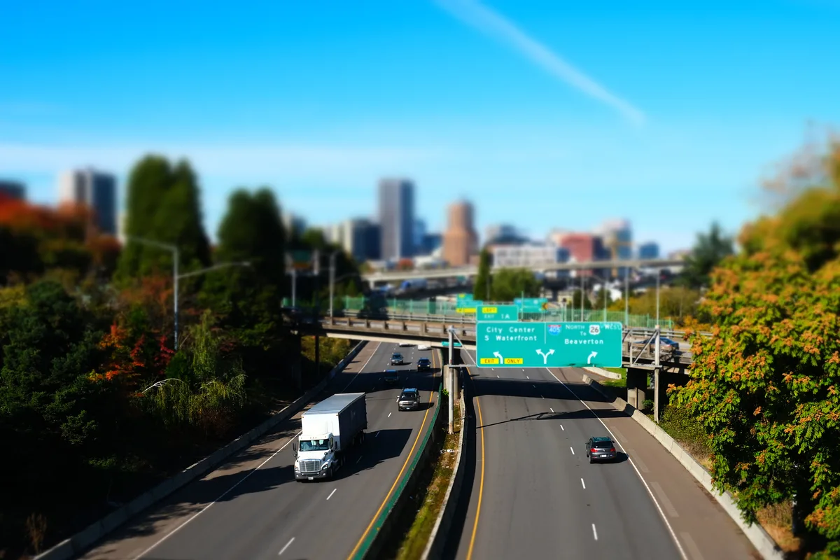 A tilt-shift photograph of Interstate 5 in South Portland, giving the scene a miniature toy-like quality. A white semi-truck and several cars travel northbound toward downtown Portland, whose skyline rises in the soft-focus background. Green highway signs direct traffic toward City Center, South Waterfront, and Beaverton via Route 26. Autumn foliage in orange, red, and green lines both sides of the freeway, and an overpass bridge and interchange infrastructure are visible in the middle distance under a vivid blue sky.